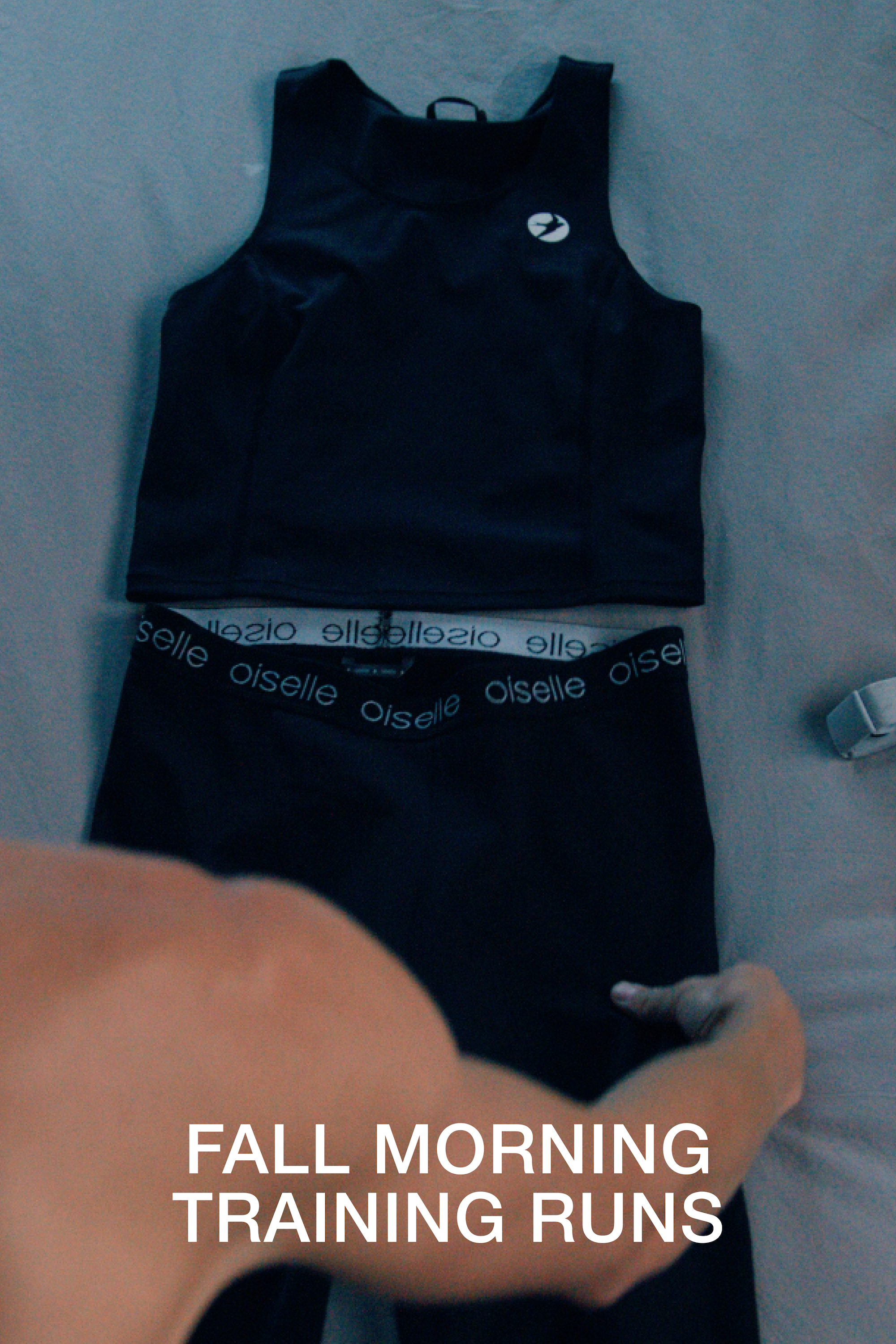 A black sleeveless athletic top and matching black shorts laid flat on a gray surface, with a visible Oiselle waistband on the shorts. A person's arm and hand are visible at the bottom, and white text at the bottom reads 'FALL MORNING TRAINING RUNS'.